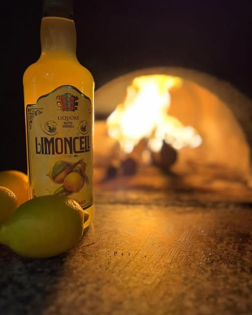 Une bouteille de limoncello et des citrons posés sur la pierre du four à bois, devant les flammes.